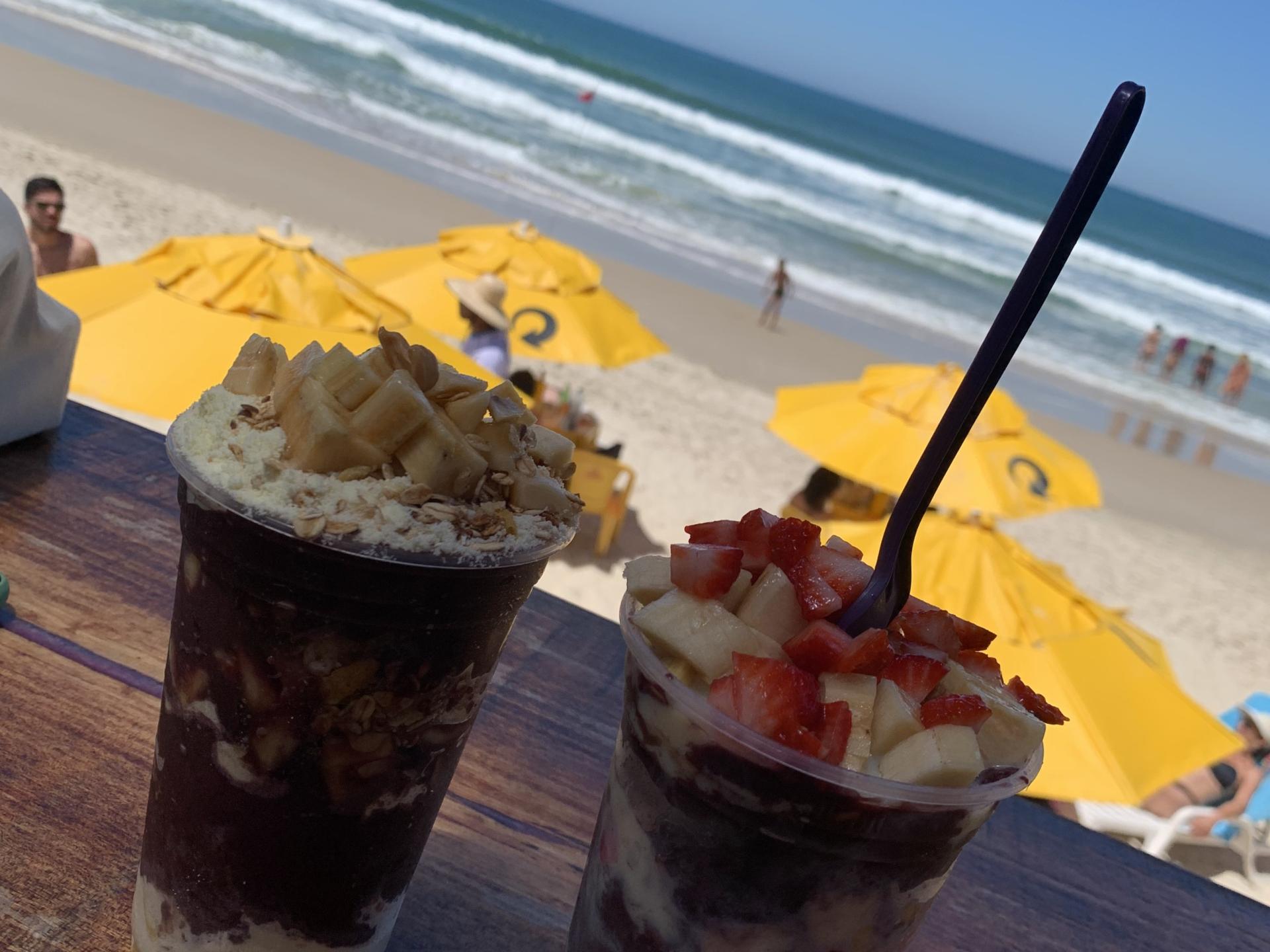 Açaí mit Früchten genießen am brasilianischen Strand Zwei Açaí-Becher mit Früchten am Strand von Brasilien vor gelben Sonnenschirmen und Meerblick