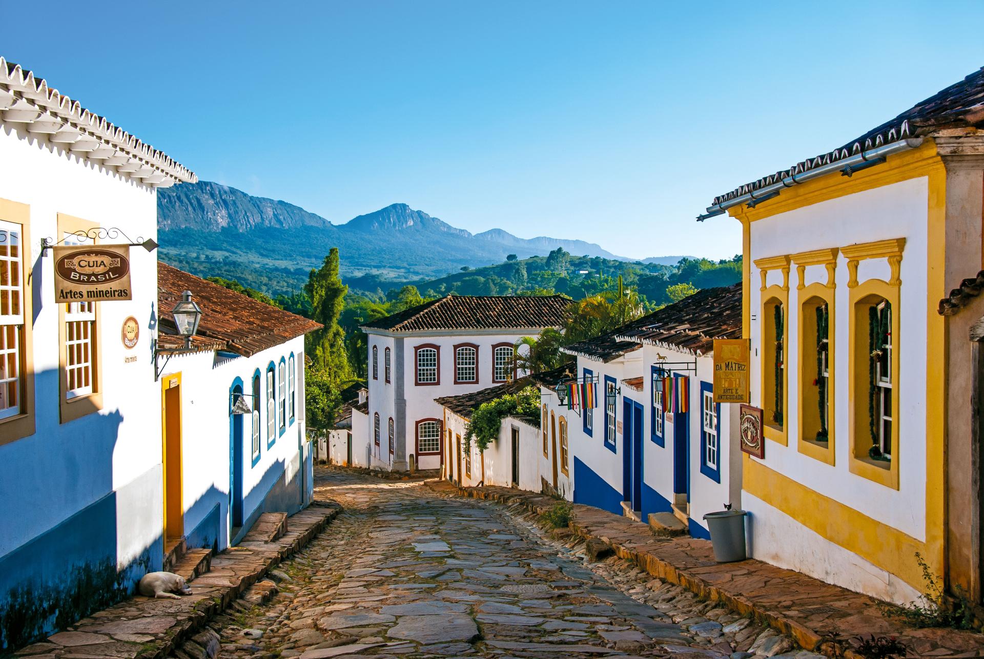 Kopfsteinpflasterstraße in Tiradentes, Minas Gerais, umgeben von bunten Kolonialhäusern mit Blick auf die Berge
